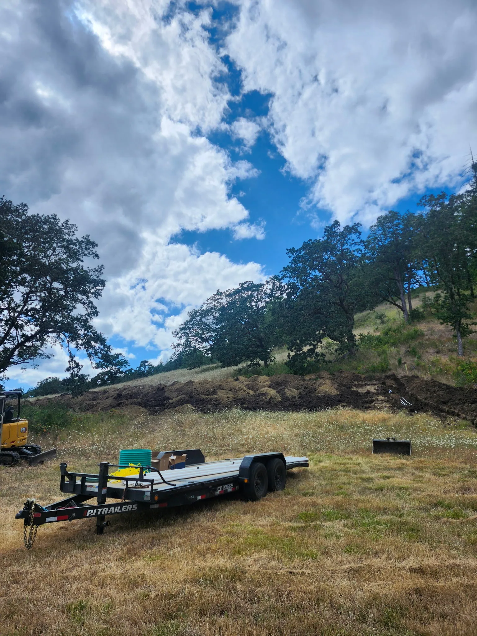 Equipment trailer parked on a rural job site below an oak hillside