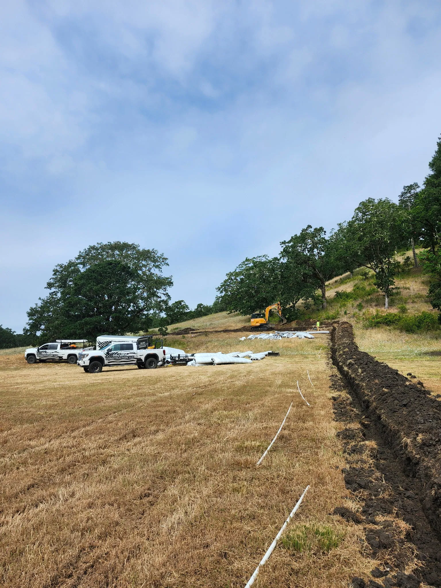 A long open trench across a dry field with pipe staged beside it