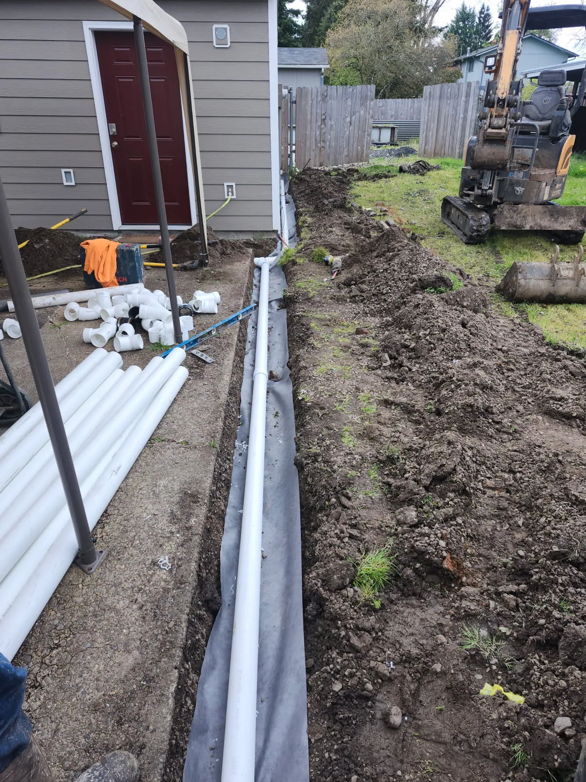 White PVC pipe staged along a side yard trench next to a house